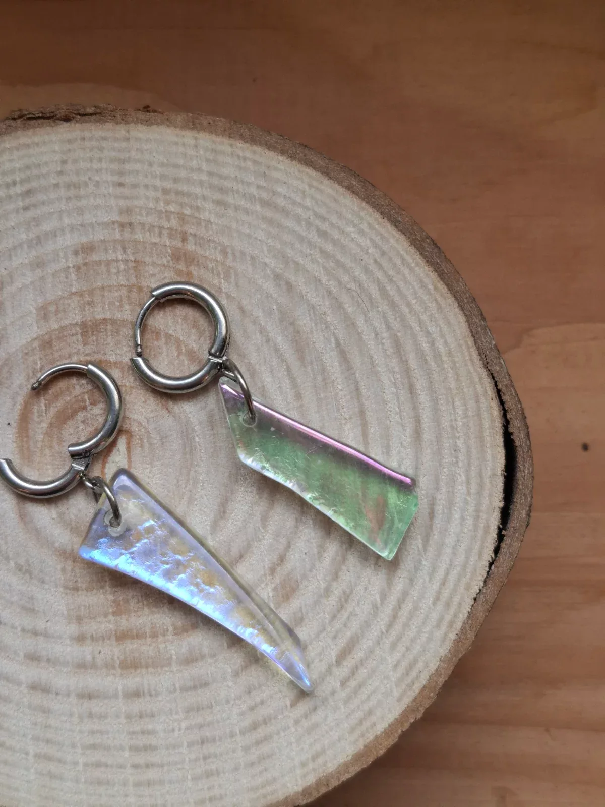 Boucles d'oreilles pendantes asymétriques en verre irisé aux reflets pastel posées sur un disque de bois.