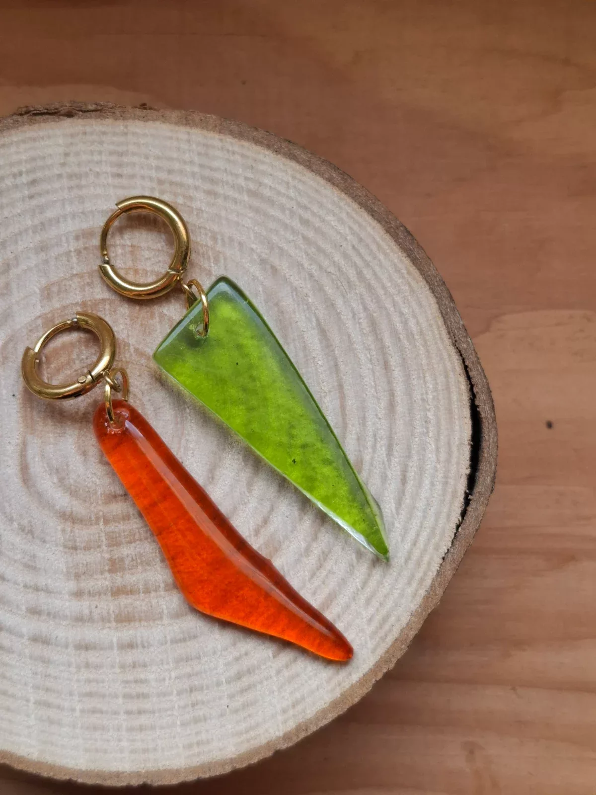Boucles d'oreilles pendantes en verre vert et orange suspendues à un présentoir doré près d'une fenêtre.