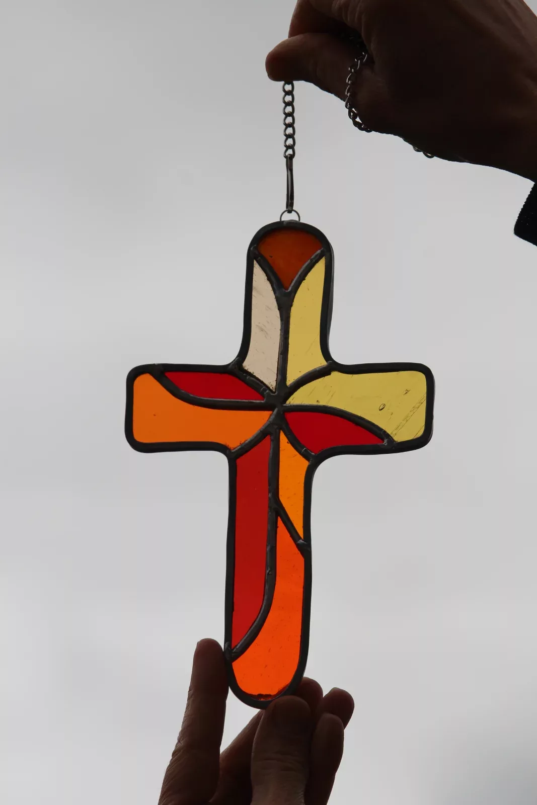 Croix en vitrail aux teintes orangées, rouges et jaunes tenue à la main en contre-jour