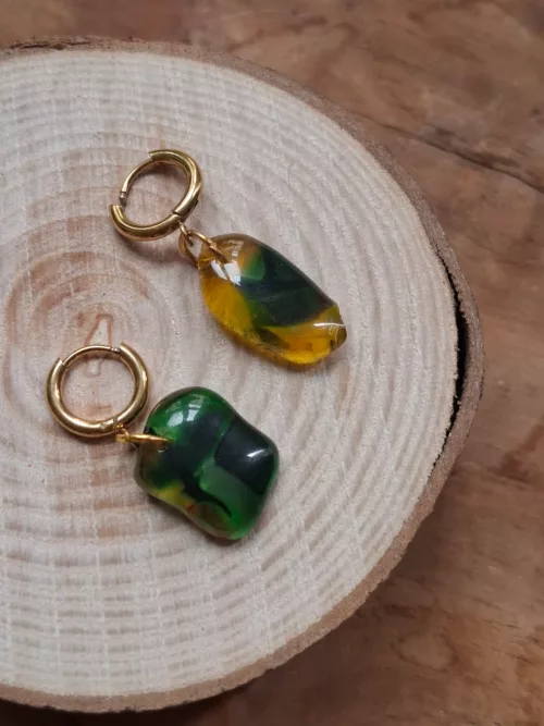 Boucles d'oreilles dorées avec pendentifs en verre vert et jaune, posées sur un disque en bois.