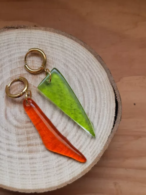 Boucles d'oreilles pendantes en verre vert et orange suspendues à un présentoir doré près d'une fenêtre.