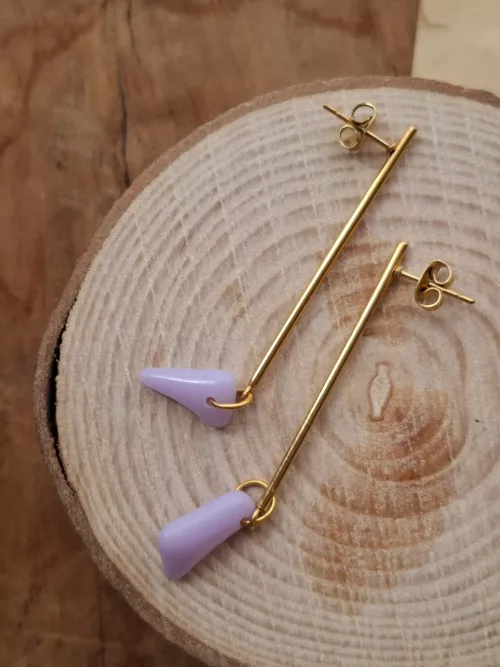 Boucles d'oreilles pendantes dorées avec pendentifs en verre violet posées sur une rondelle de bois.