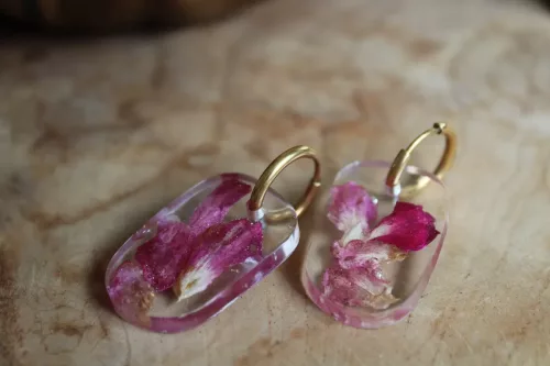 Boucles d'oreilles ovales transparentes avec pétales roses et anneaux dorés posées sur du bois.