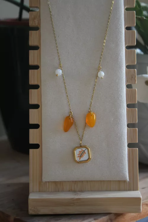 Collier doré à pendentif carré végétal et pampilles orange présenté sur un porte-bijou en bois