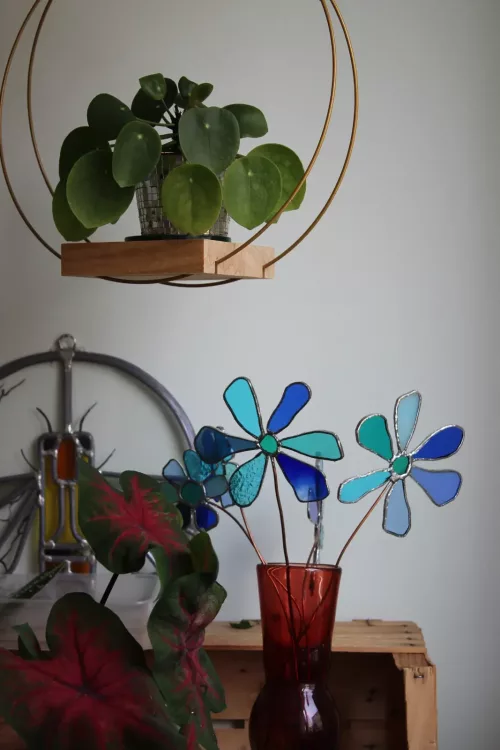 Deux fleurs en vitrail bleu dans un vase rouge, sous une plante verte suspendue.